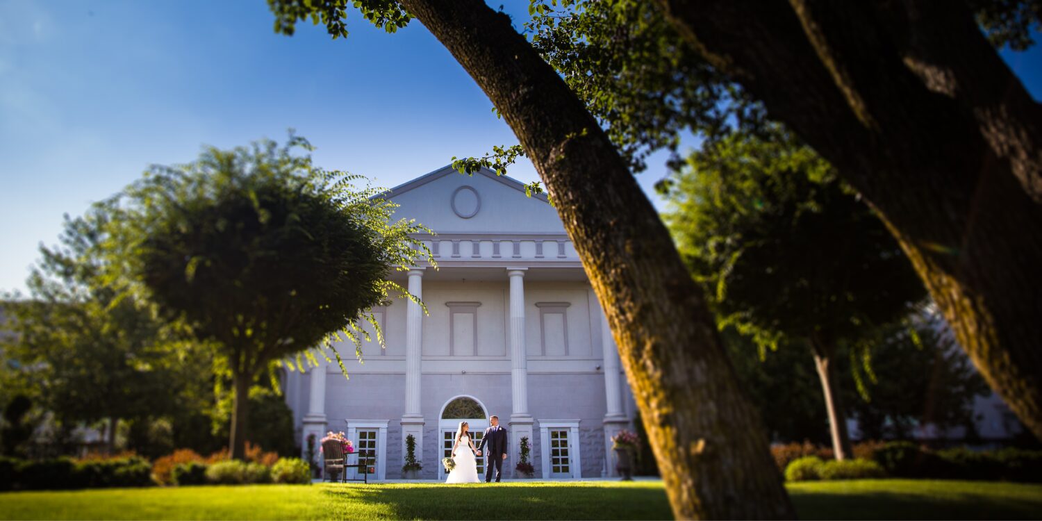 palace at Somerset wedding photographer, Rhinehart Photography, captures this landscape image of the bride and groom as they walk together outside of the palace at Somerset Park in New Jersey 