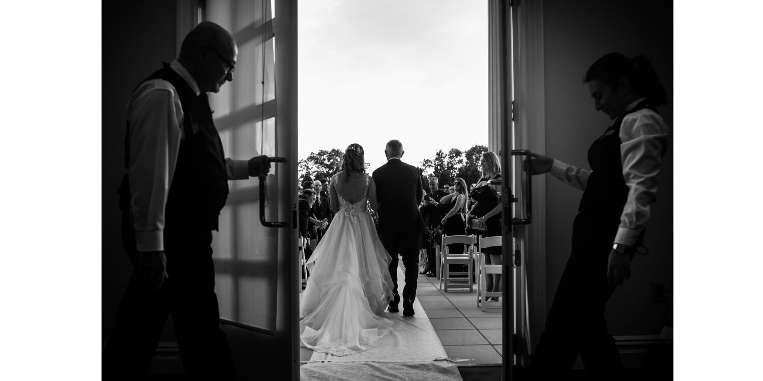 black and white image of the bride and her father as they walk down the aisle together while the palace at Somerset Park opens the doors for them during this outdoor wedding ceremony 