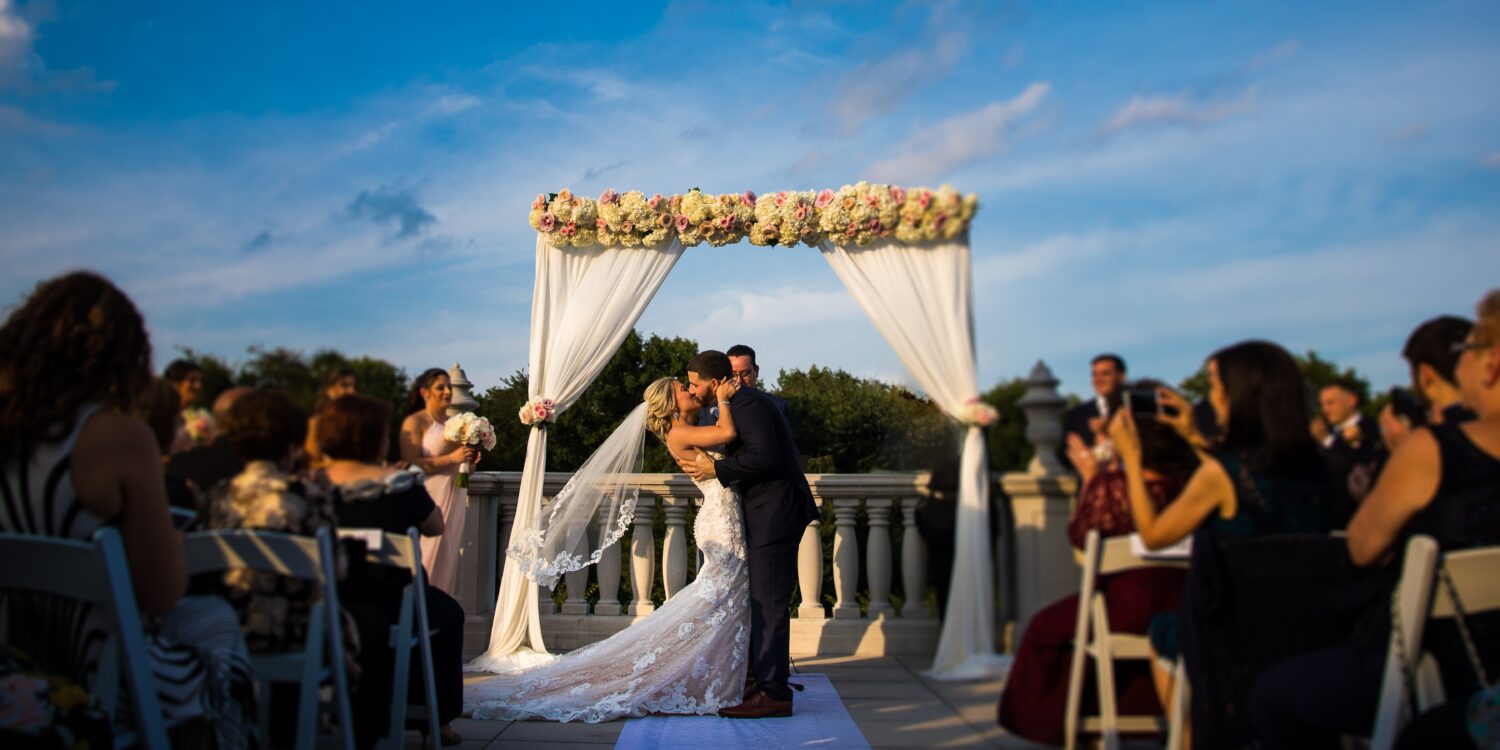 vibrant, colorful image of the bride and groom as they kiss after their outdoor wedding ceremony with a vibrant blue sky in the background behind them during this palace at Somerset Park wedding ceremony 