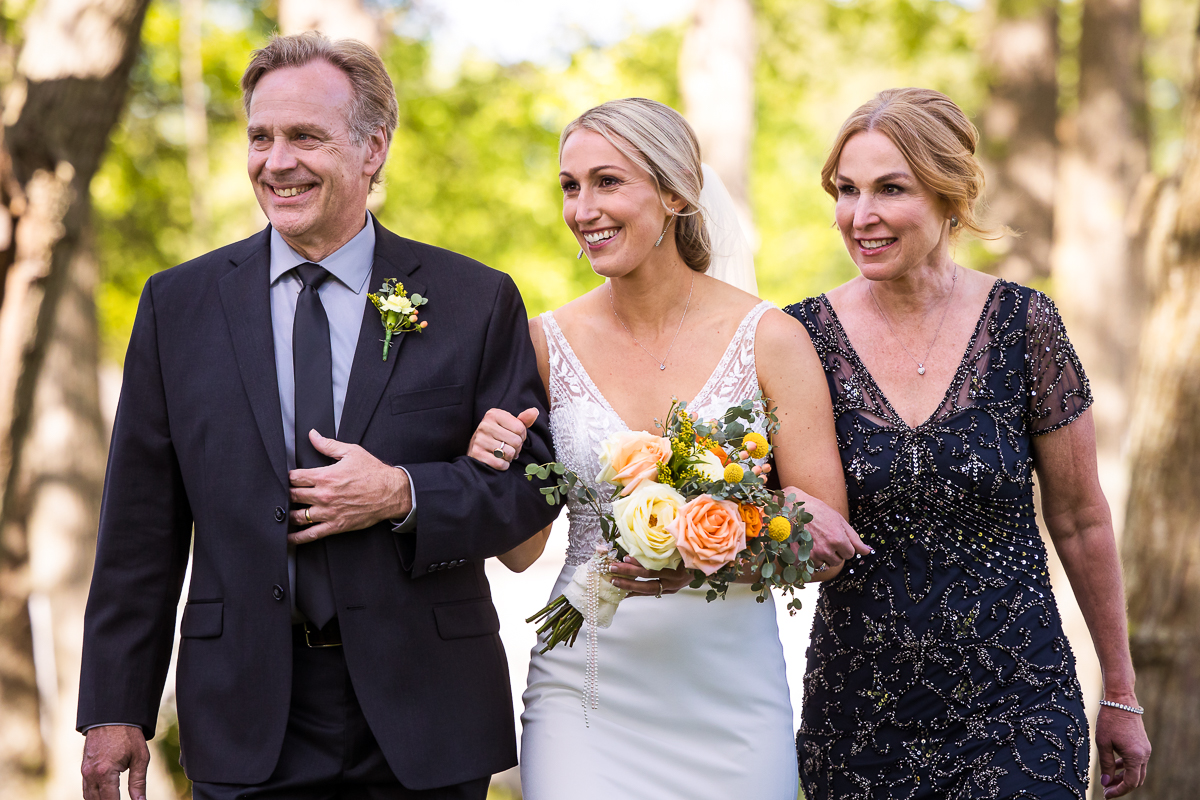 bride carrying bouquet being walked down the aisle by both parents outdoor ceremony