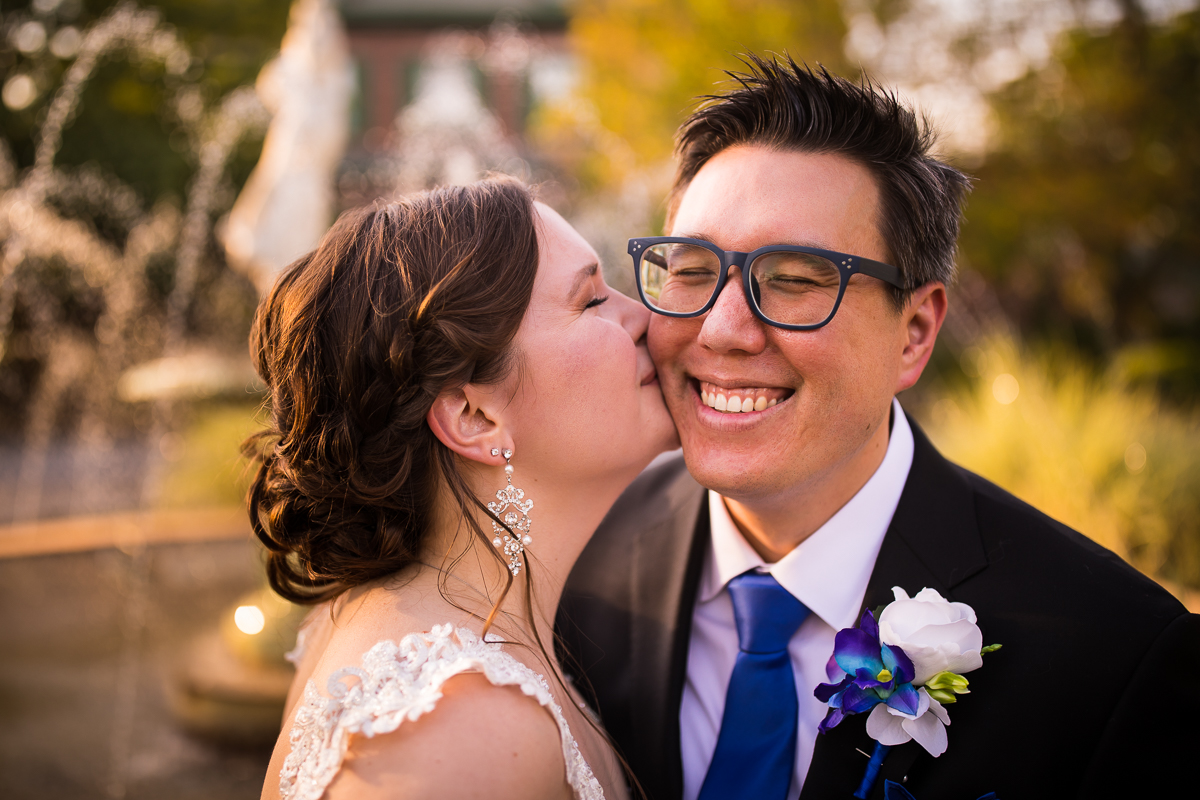 bride giving groom wearing glasses kiss on cheek after wedding ceremony groom wearing blue tie and flowers
