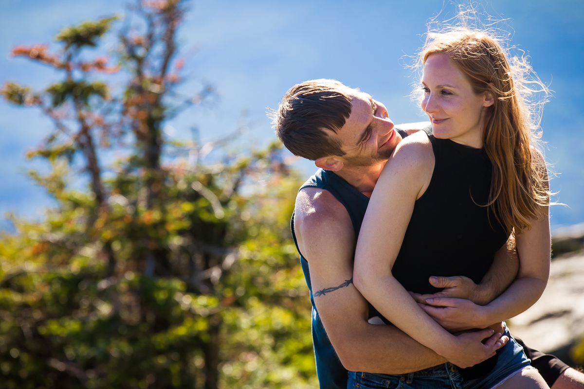 guy hugging girl from behind smiling at each other wearing black tank tops on top of mountain with trees in the background