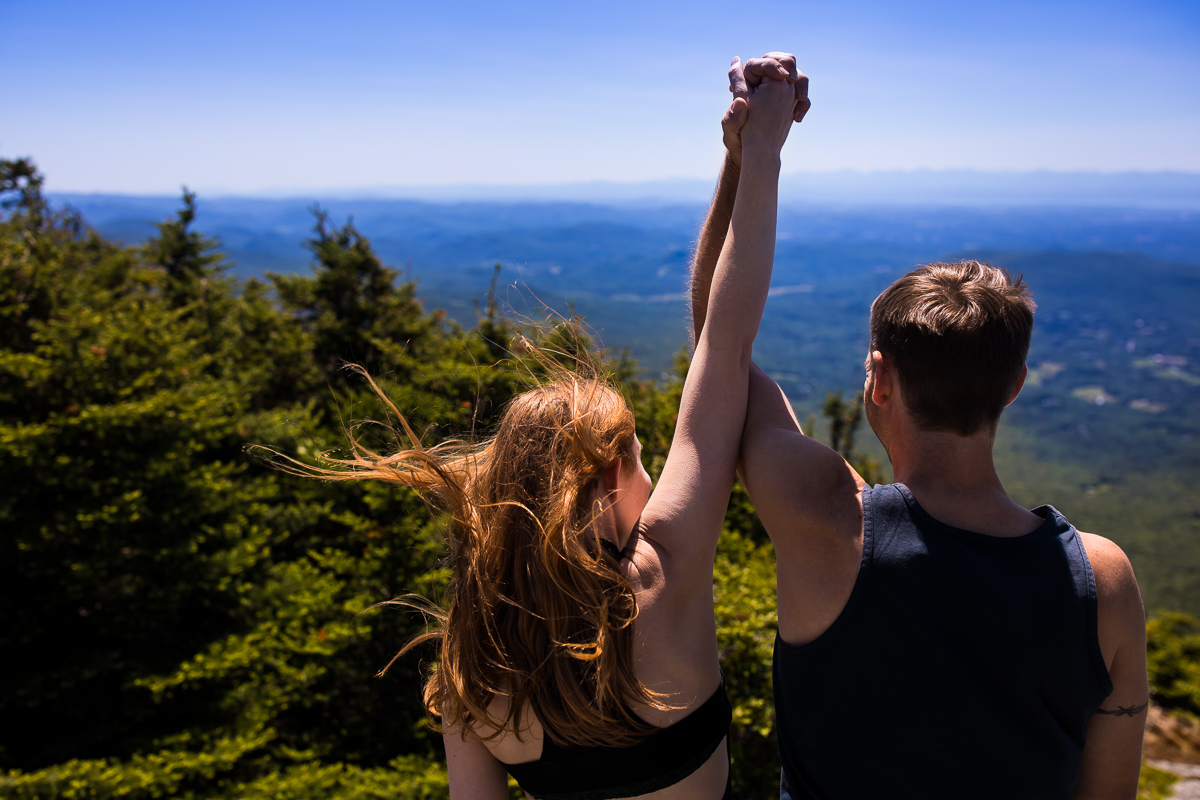 couple holding hands up in the air at the top of the summit of mount Mansfield in Burlington vt with trees and ground in the distance