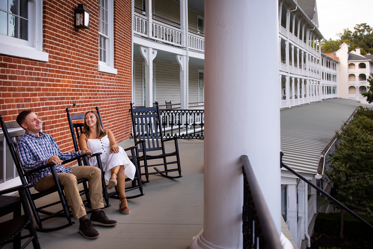 best award winning omni Bedford Springs photographer unique authentic couple sitting on rocking chairs in front of brick wall outside omni resort girl wearing white dress guy wearing blue plaid shirt 