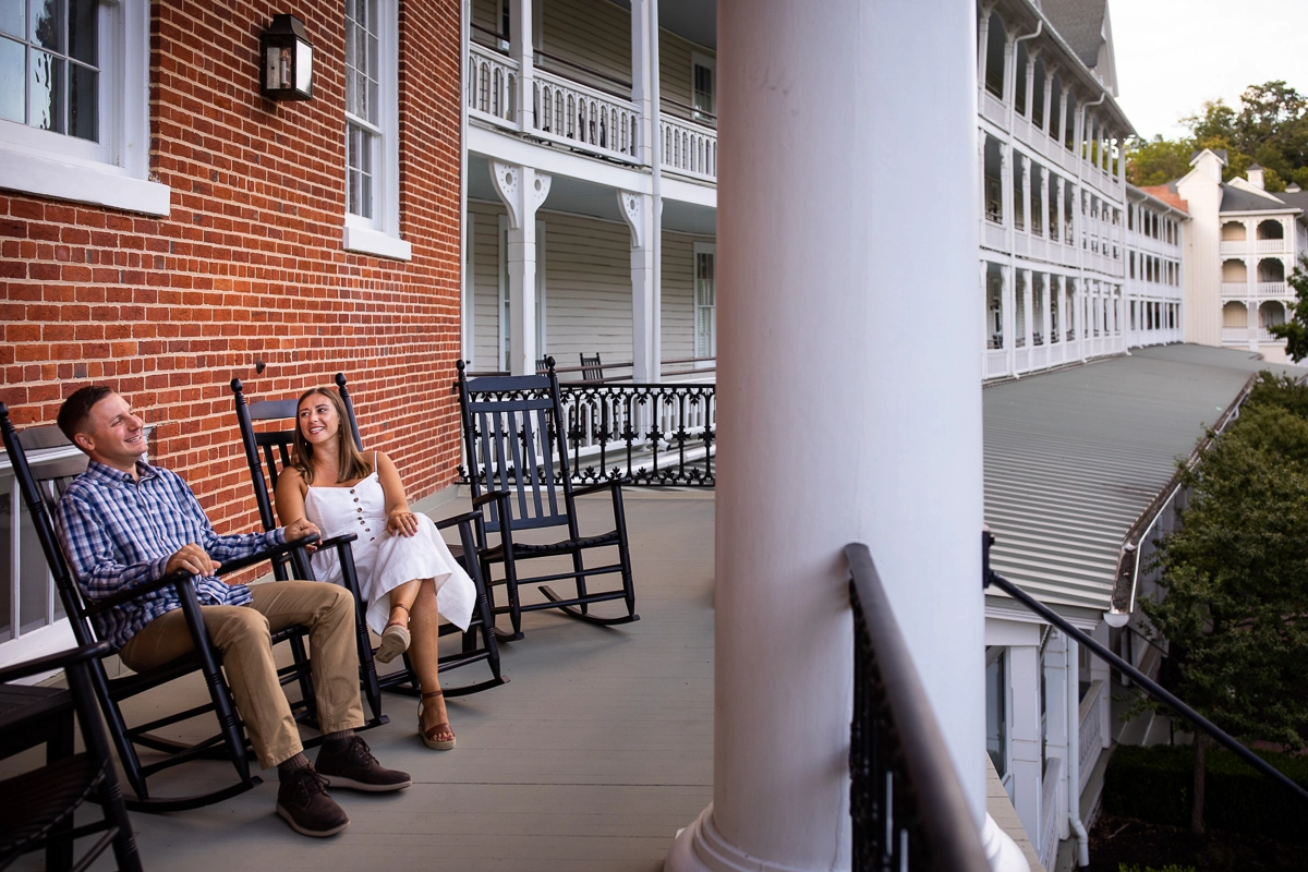 best award winning omni Bedford Springs photographer unique authentic couple sitting on rocking chairs in front of brick wall outside omni resort girl wearing white dress guy wearing blue plaid shirt