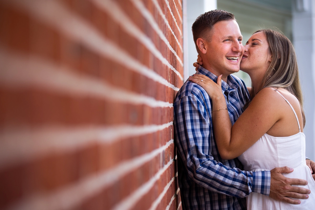 vibrant authentic award winning engagement photographer couple leaning against brick wall girl wearing white dress kissing guy on cheek with arms around each other