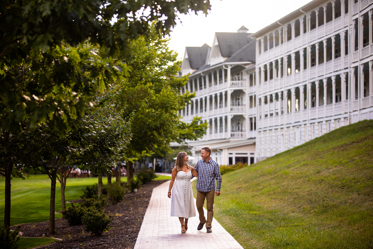 omni Bedford Springs engagement photographer couple walking along tree lined path in front of omni resort hand in hand looking at each other smiling