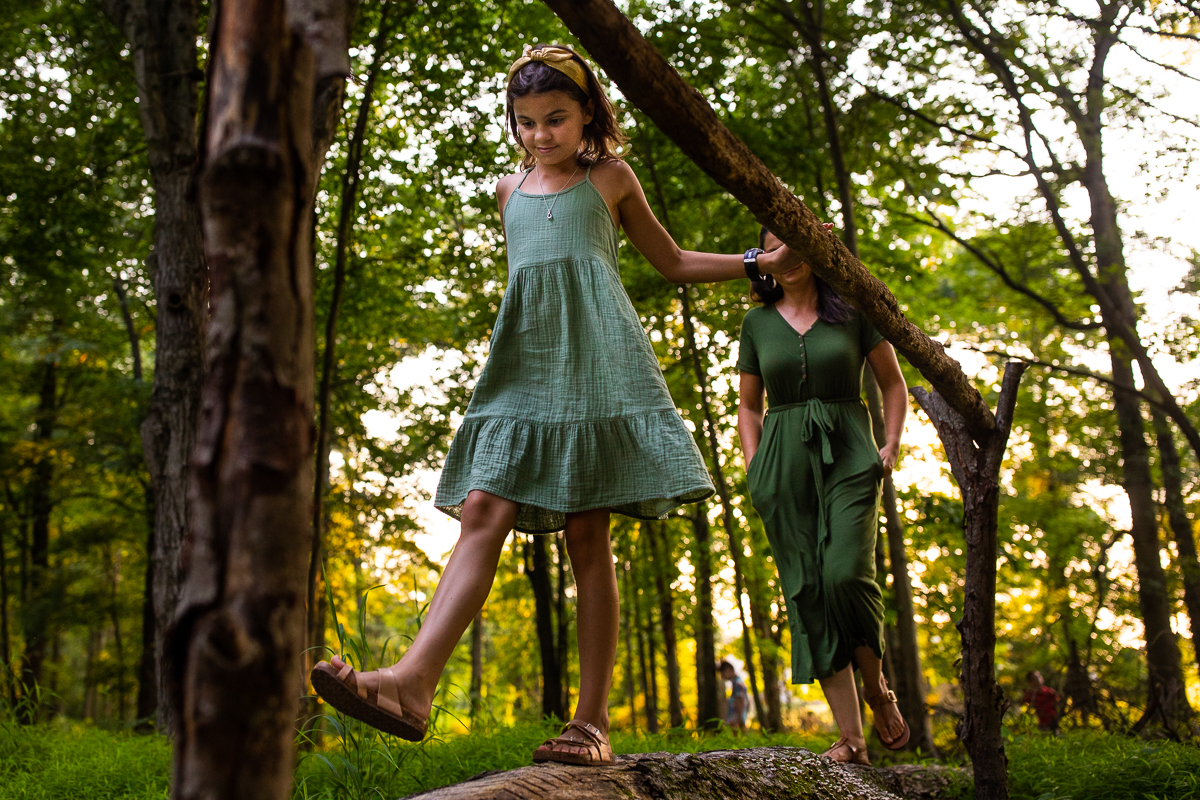 vibrant candid authentic award winning family photographer farm lifestyle girl wearing green dress walking on log while mom follows behind in middle of forest