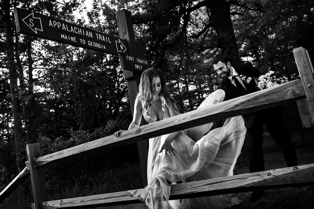Appalachian trail hiking photographer black and white photo of bride climbing over fence while groom helps hold dress 