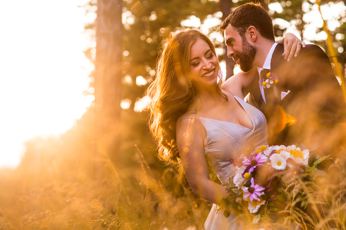 best award winning central pa sunset photographer couple laughs with each other as bride puts arm around groom holding bouquet in woods