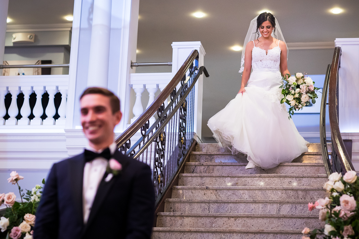 arts ballroom Philadelphia wedding best wedding photographer bride walks down stairs during first look groom in foreground smiling