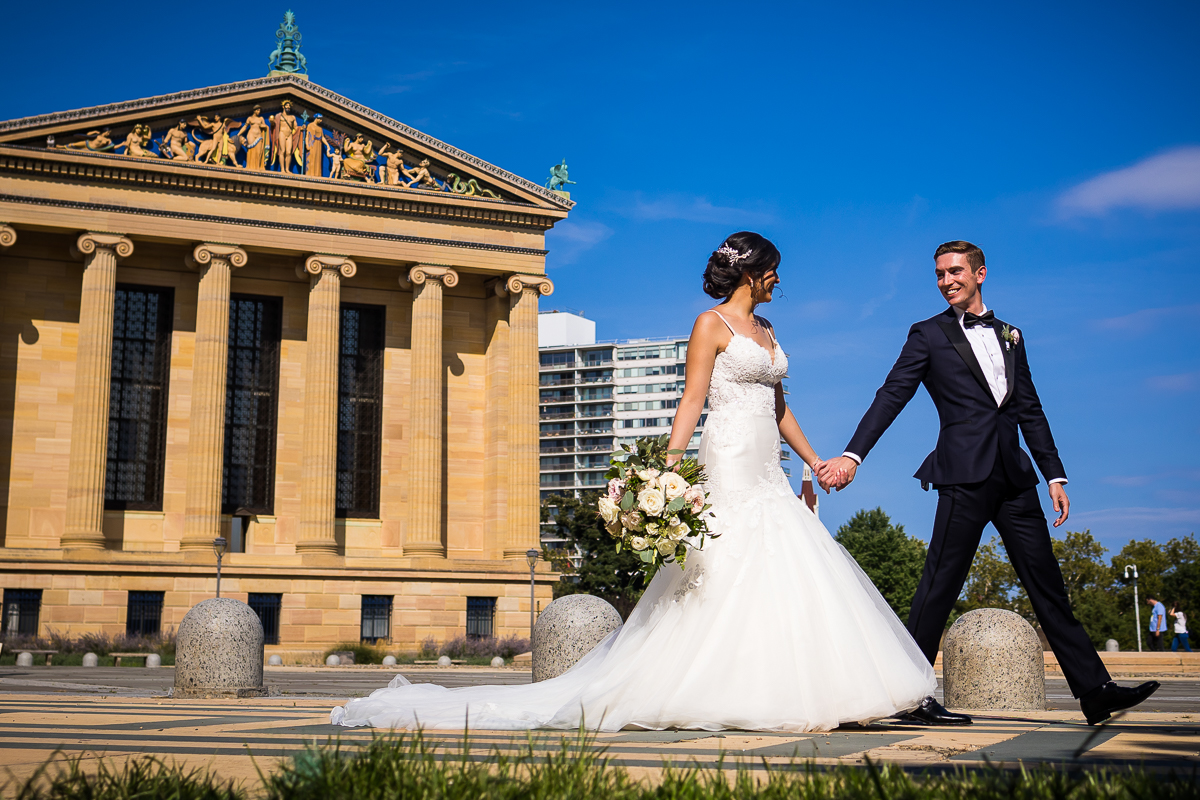 vibrant creative downtown Philadelphia wedding photographer bride and groom smile and walk hand in hand with each other in front of Philadelphia museum of art