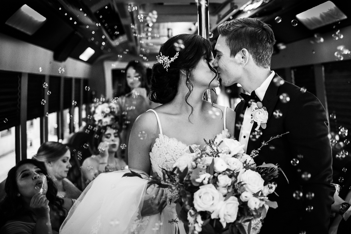 best award winning wedding photographer black and white photo of bride and groom kissing on party bus while wedding party blows bubbles around them