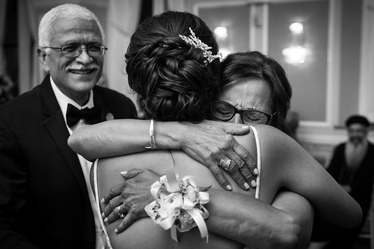 emotional authentic wedding photographer black and white photo of mom hugging bride on wedding day while dad smiles in background