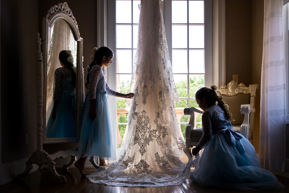 creative artistic central pa wedding photographer two girls in blue dresses look at bride's wedding dress hanging in front of window