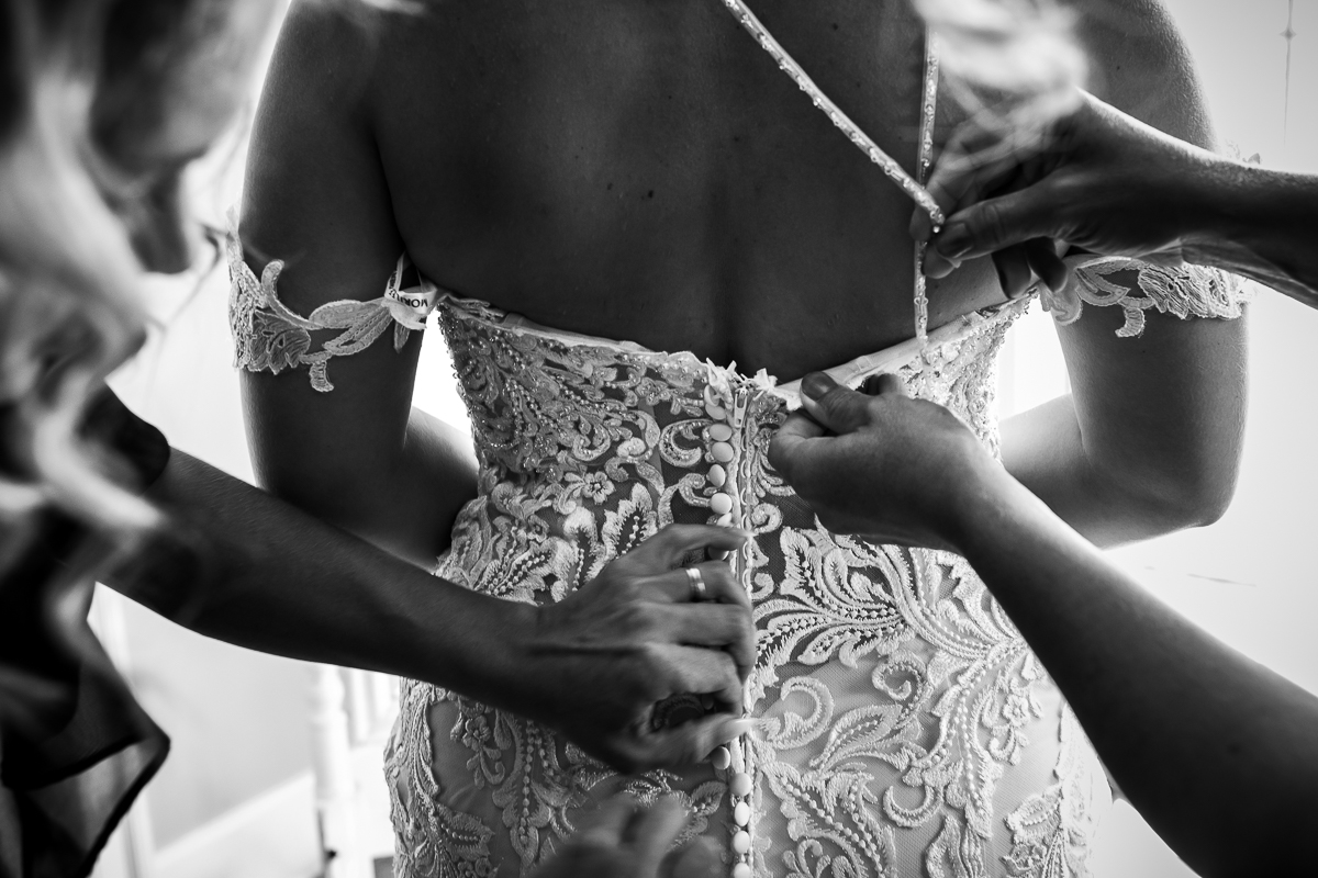 black and white photo of hands helping button the bride's wedding dress intricate lace detail and buttons thin beaded straps