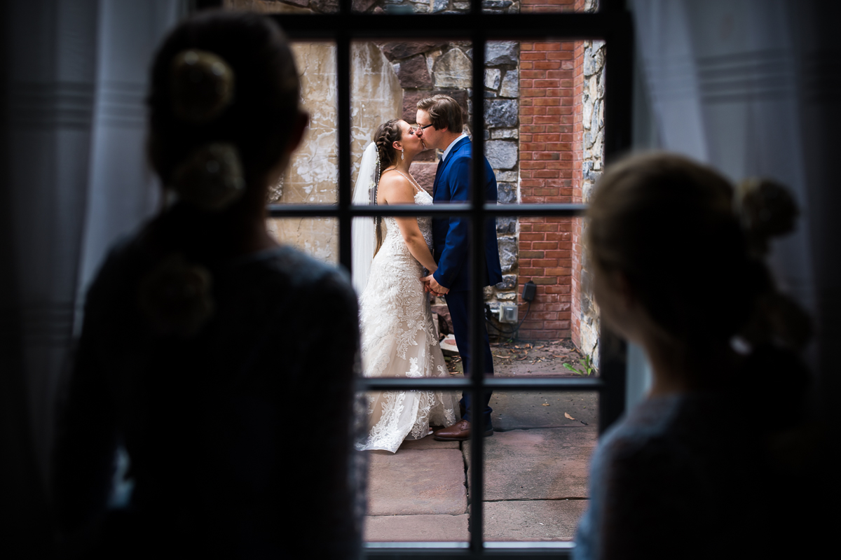 bride and groom share kiss best award winning historic acres of Hershey wedding photographer while girls look on through the window 