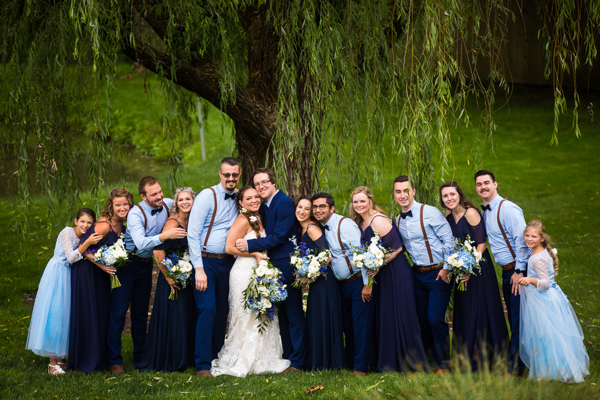 bride and groom with large wedding party dressed in blue stand outside under willow tree at historic acres of Hershey