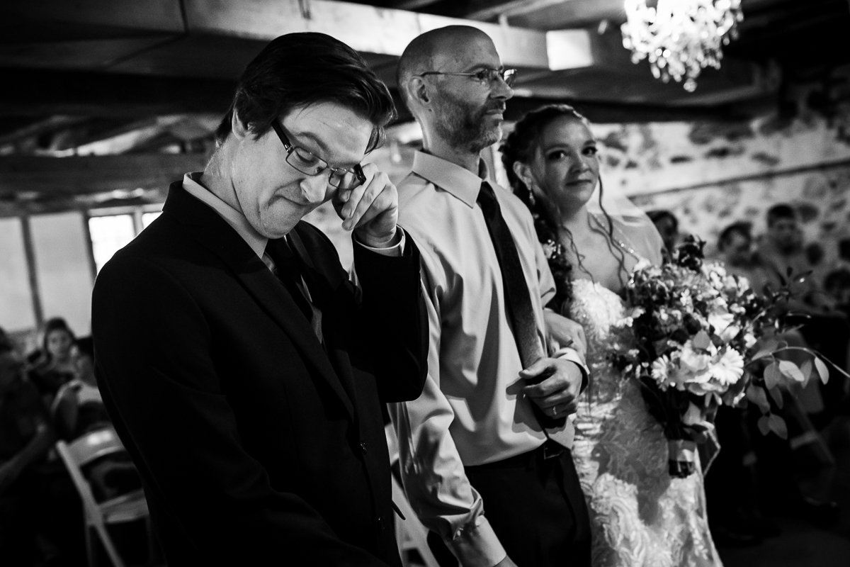 emotional candid central pa wedding photographer black and white photo of groom wiping tear from eye while father of bride escorts bride down the aisle