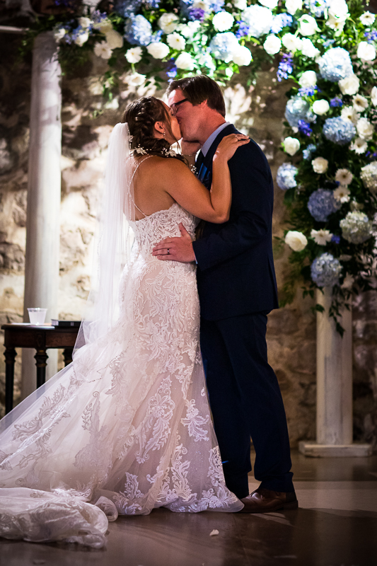 bride and groom share first kiss with blue and white flowers behind them indoors during historic acres of Hershey wedding ceremony 