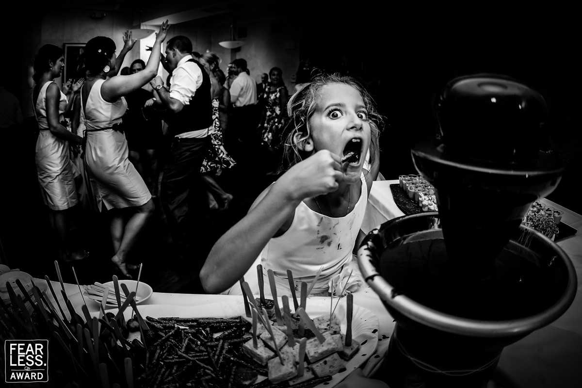 This Fearless Photographers Award winning photo is a black and white photograph of a young girl eating from a fountain of chocolate while everyone is dancing around in the background ranking lisa rhinehart as one of the Best wedding photographers in the USA