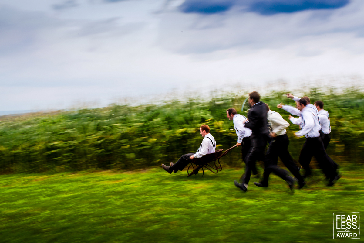 Vibrant, creative groomsmen group photo of them pushing the groom in a wheelbarrow running through a field for another Fearless Photographer award for Lisa Rhinehart one of the Best wedding photographers in the USA and ofcoaurse also the best wedding photographers in pa Pennsylvania 