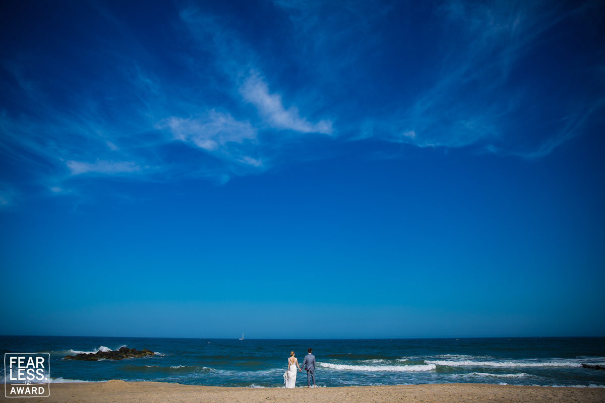 Colorful, vibrant photo of the couple standing on the beach out by the ocean with a bright blue sky received a Fearless Photographer Award for Rhinehart Photography