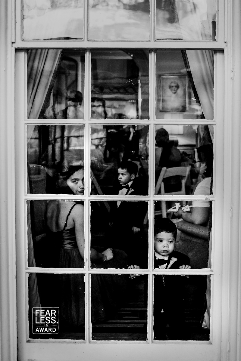 Black and white creative image taking from outside of the venue looking in through the window with a young boy and bridesmaid looking out the window