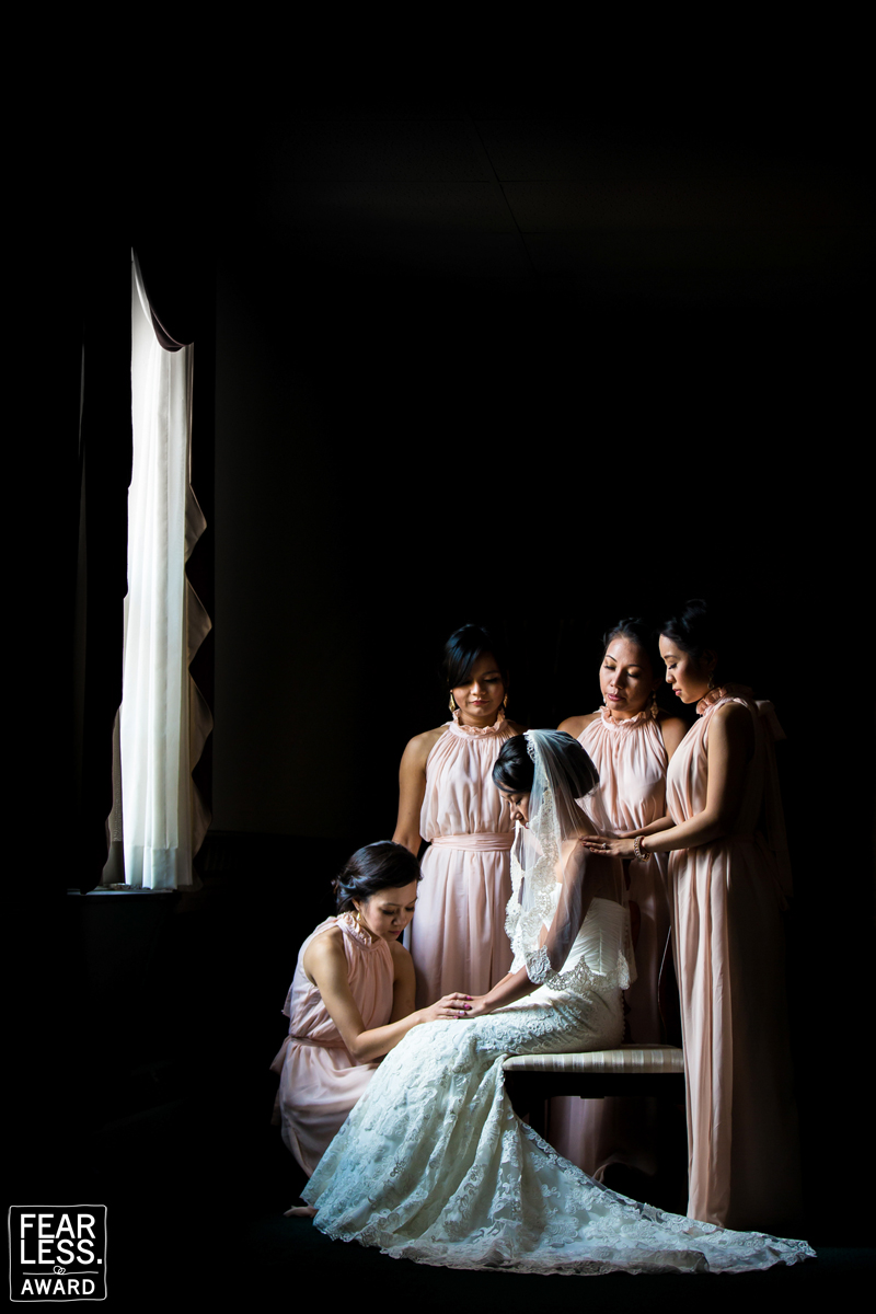 Dark, dramatic photo of the bridesmaid standing around the bride with their heads down and the light from the window shining in on them best wedding photographers in dc 