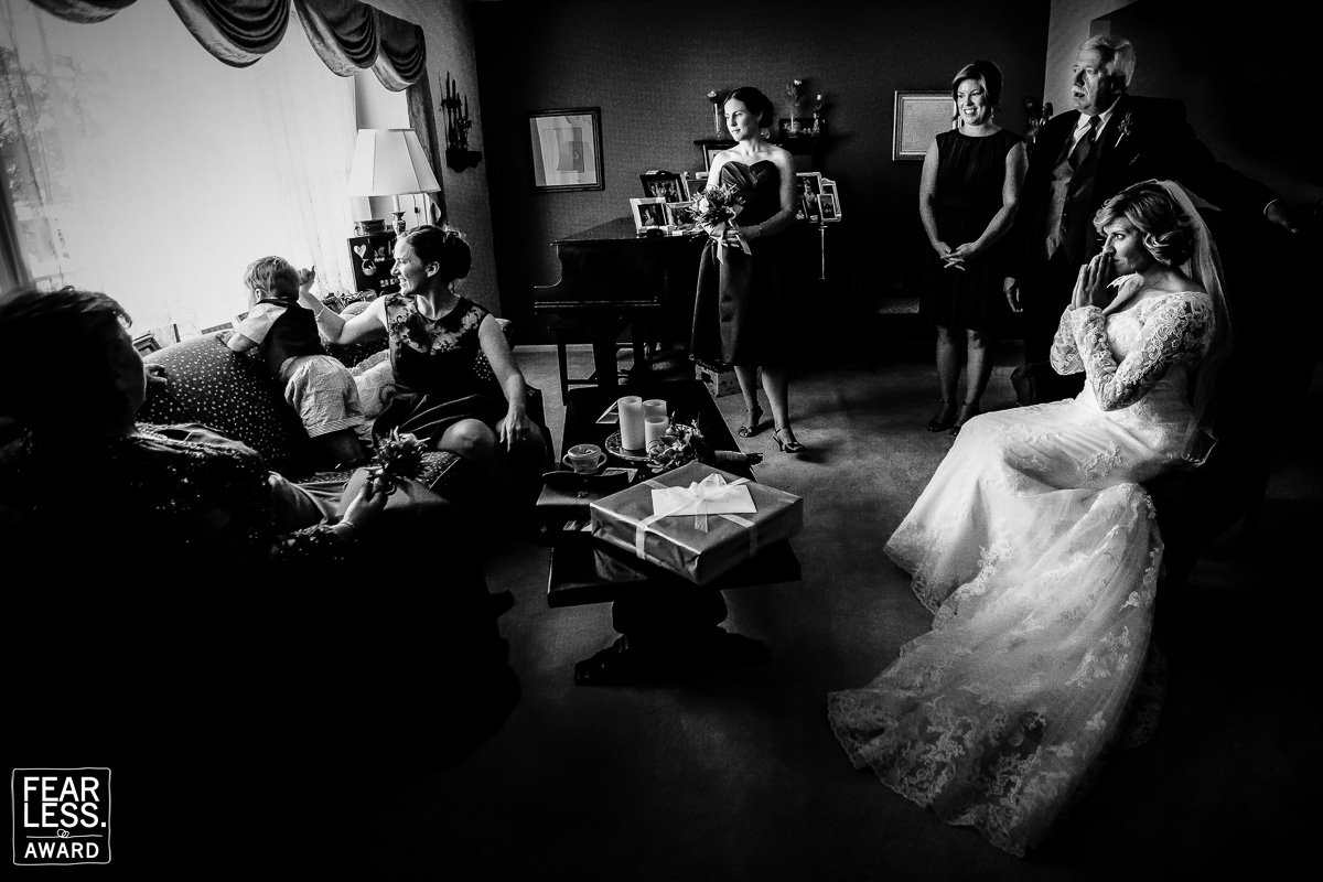 Black and white image of the bride, family members and the wedding party inside looking at the baby climbing on the couch towards the window
