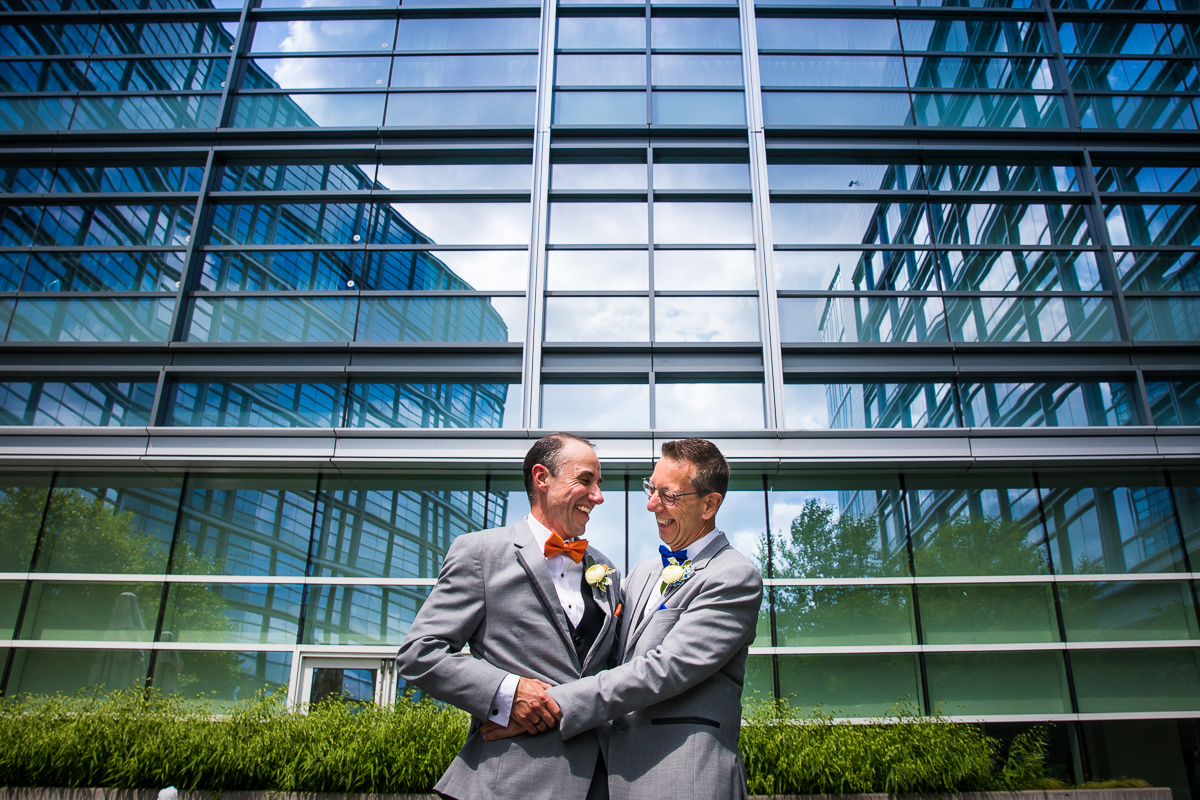 best award winning DC photographer two grooms hold each other during first look with building architecture behind them wearing gray suits smiling at each other 