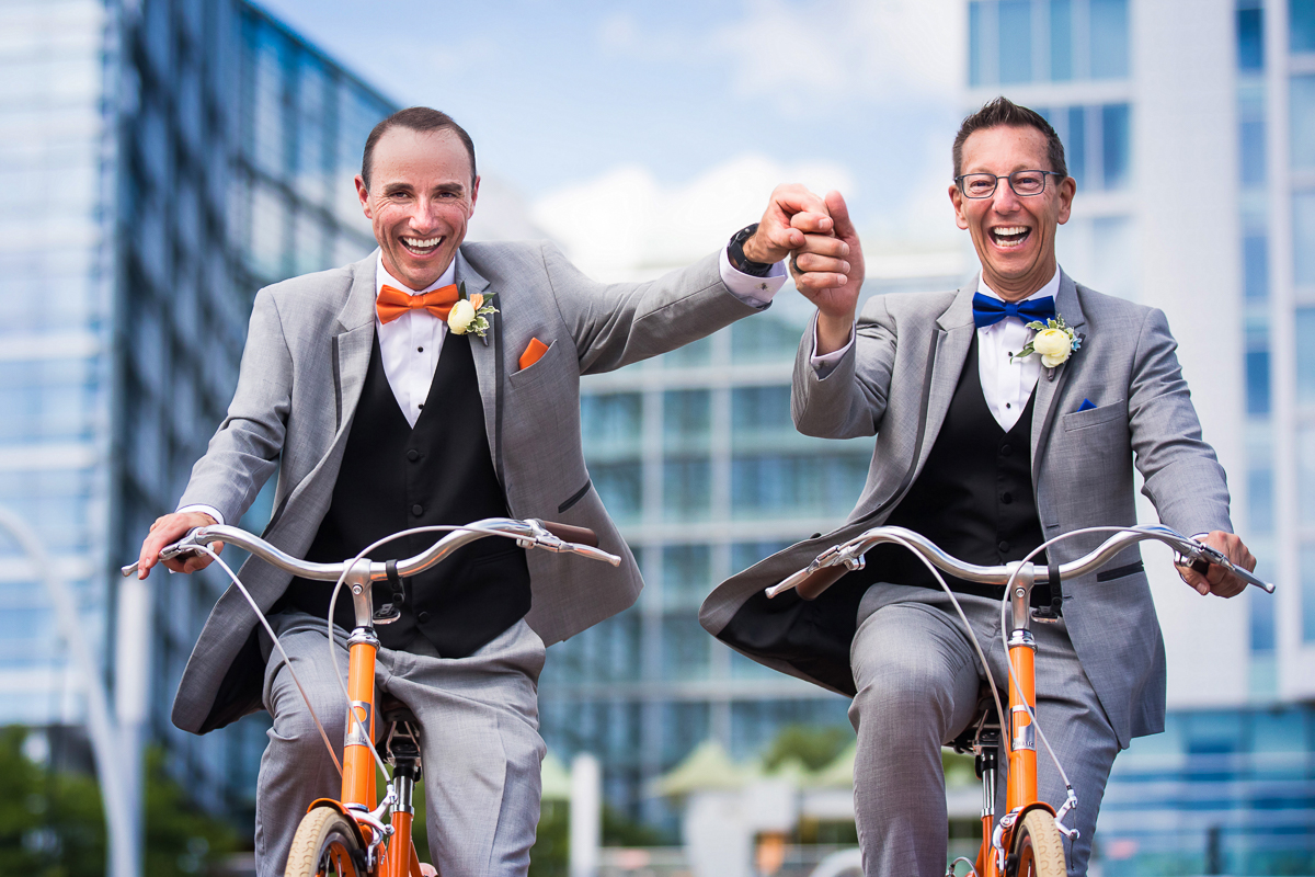 vibrant creative colorful best award winning photographer two grooms holding hands while riding orange bikes smiling at the camera outside DC wharf hotel best award winning national arboretum wedding photographer