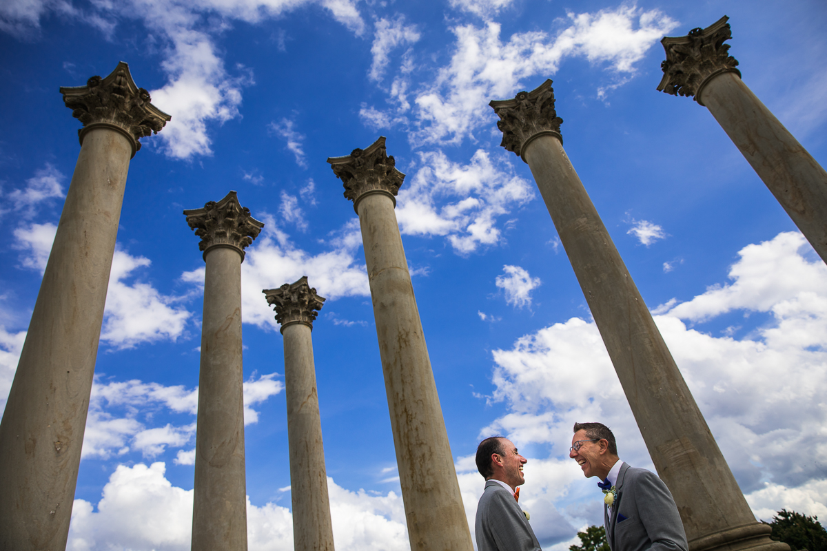 vibrant authentic award winning DC wedding photographer National Capitol Columns two grooms stand underneath columns smiling at each other with vibrant blue cloudy sky in background