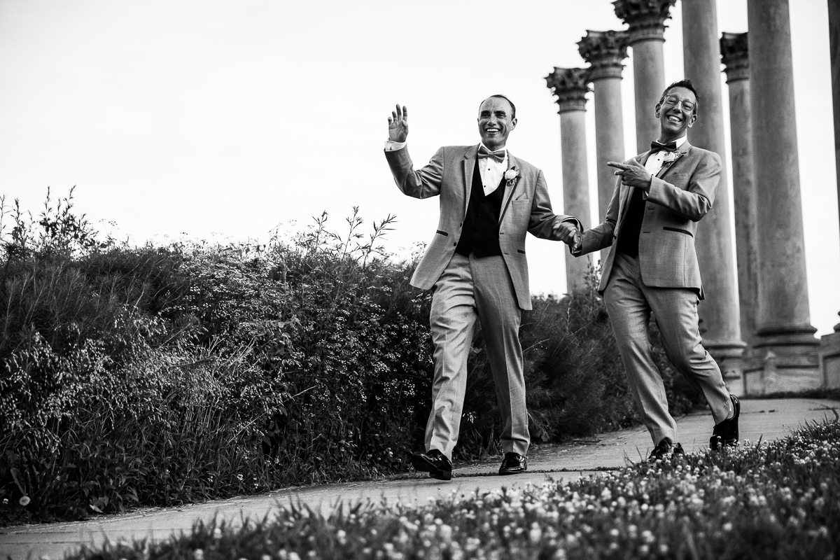 black and white photo of two grooms holding hands walking and waving at the National Capitol Columns Washington DC best award winning wedding photographer