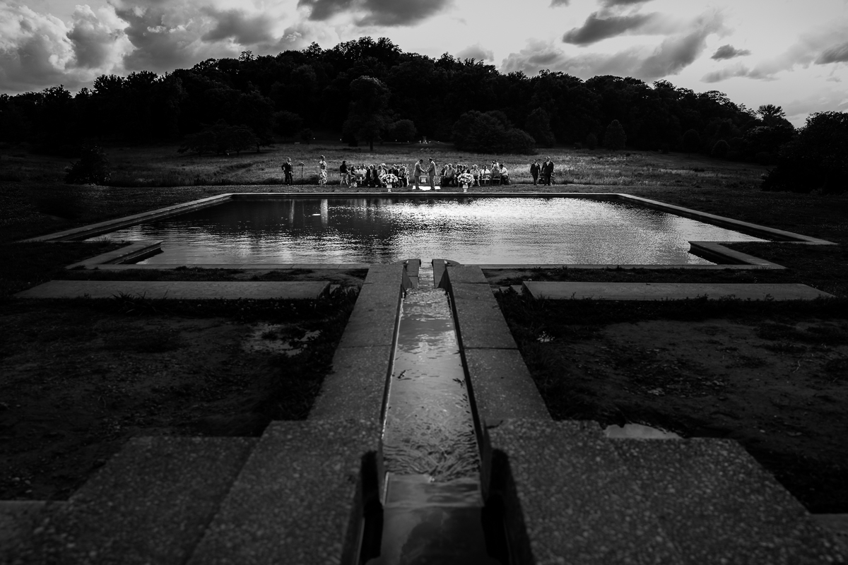 black and white photo of pool and dramatic skies at national arboretum wedding two grooms holding hands with guest in background during ceremony