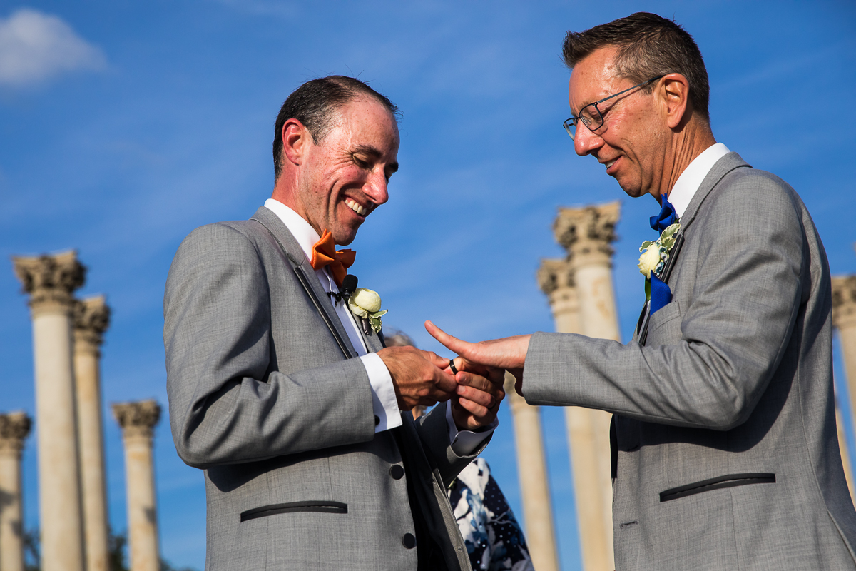 colorful authentic candid best DC wedding photographer groom places ring on groom finger during wedding vows at national arboretum wedding with bright blue Skies behind
