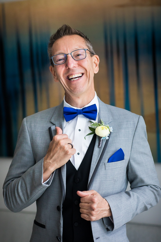 wharf dc Canopy Hilton wedding photographer groom with glasses holding jacket wearing gray suit with blue bowtie smiling at camera during getting ready portraits