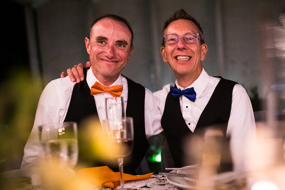 national arboretum wedding reception speeches two grooms smile with arms around each other while listening at their table wearing orange and blue bowties authentic candid wedding photographer