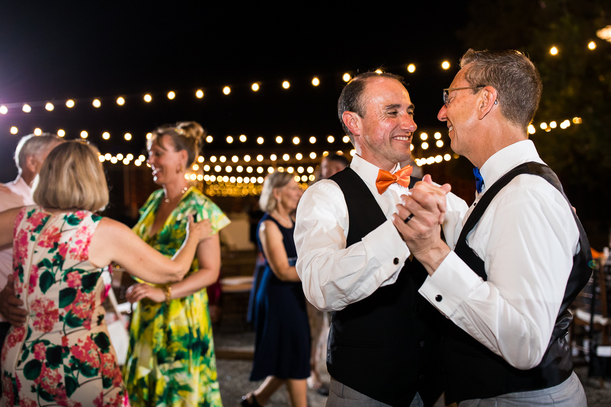 best national arboretum wedding photographer two grooms holding hands dancing together during wedding reception on patio with guests behind them