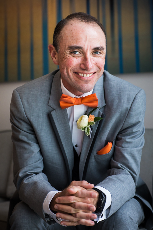 best award winning wedding photographer dc groom sitting on couch wearing gray suit and orange bow tie getting ready at Hilton canopy wharf hotel dc
