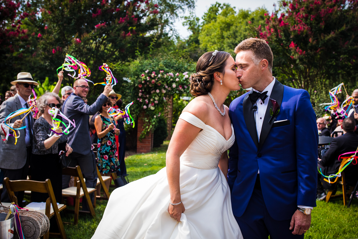 river farm wedding Alexandria Virginia couple kiss after walking down the aisle as guests wave colorful streamers during outdoor ceremony