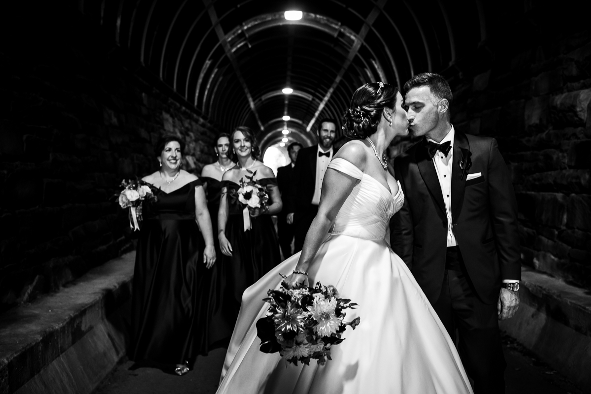 black and white photo of bride and groom kissing with wedding party walking behind them in Wilkes street tunnel best dc wedding photographer