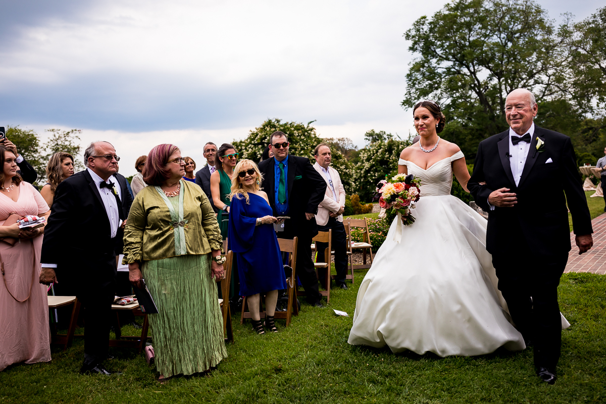 authentic emotional wedding photographer bride walks down aisle with father as guests look on during outdoor wedding ceremony at river farm