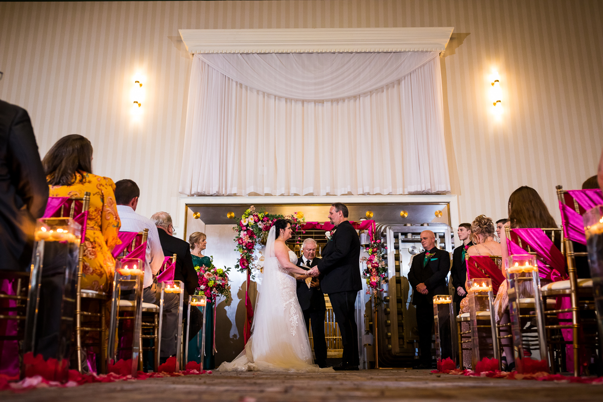 gettysburg hotel wedding ceremony bride and groom hold hands while officiant stands behind pink flowers and sashes around chairs candles romantic vibe