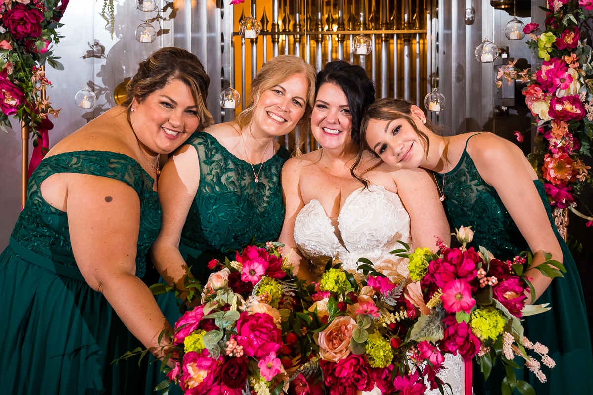 bride with bridesmaids holding pink and green flower bouquets smiling during cocktail hour in front of wall with floating candles