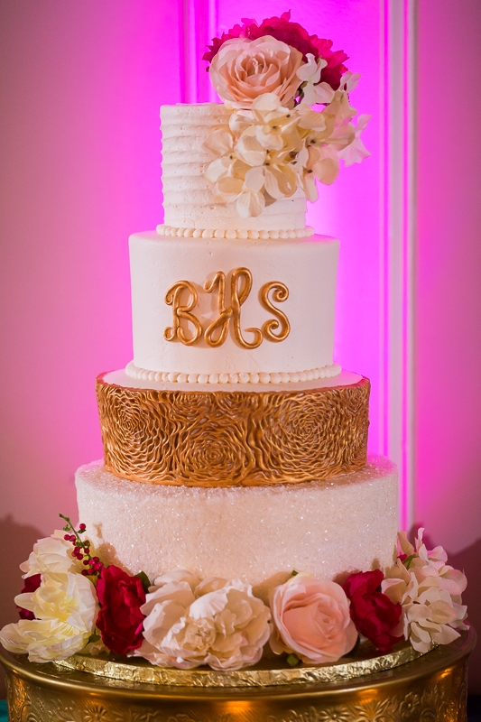 white and gold wedding cake topped with pink and white flowers monogrammed with pink uplighting behind gettysburg hotel reception