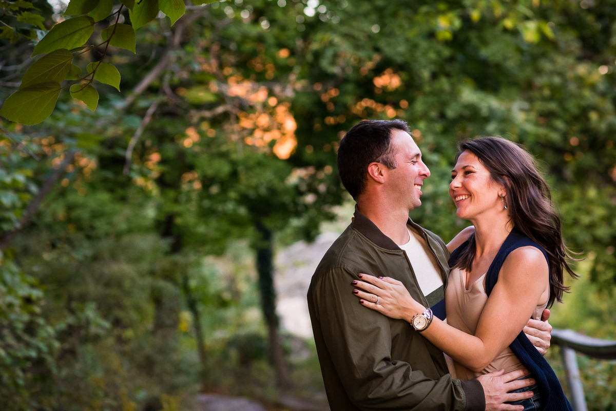fall engagement session couple standing next to river smiling and looking at each other with trees behind