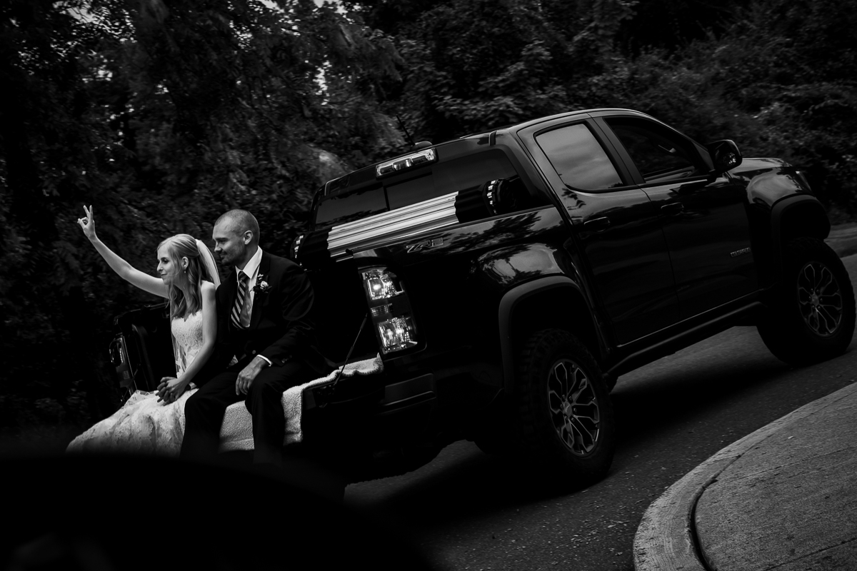 black and white photo of bride and groom smiling bride holding up peace sign in back of groom's truck