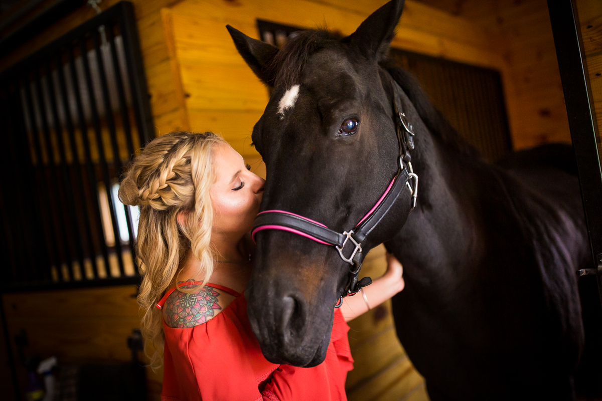 black horse in a barn stall being kissed on the nose by a blonde woman with a red shirt during her engagement photography session