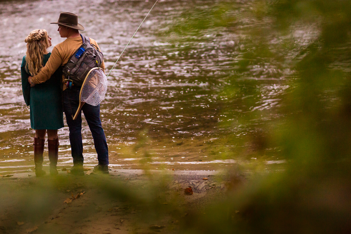 horseback riding and fly fishing engagement session couple standing by water holding fly fishing gear looking at each other